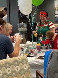 An elf holds up a balloon next to some family members at the table